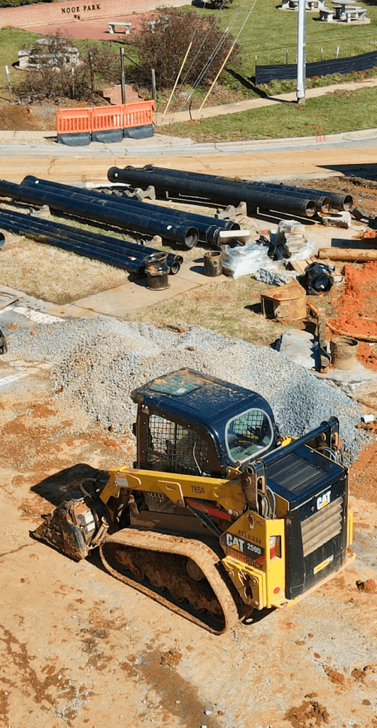 Construction site with a small CAT loader, pipes, and materials laid out near a park.