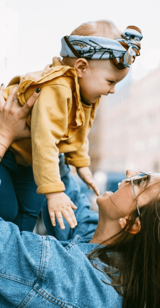 Joyful mother in a denim jacket holding her laughing toddler who wears a stylish headband and mustard-colored outfit.