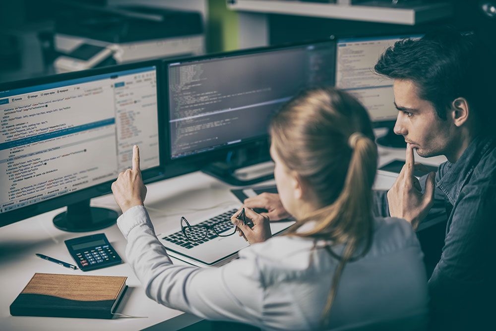 Two software developers working on code together at a multi-monitor workstation.