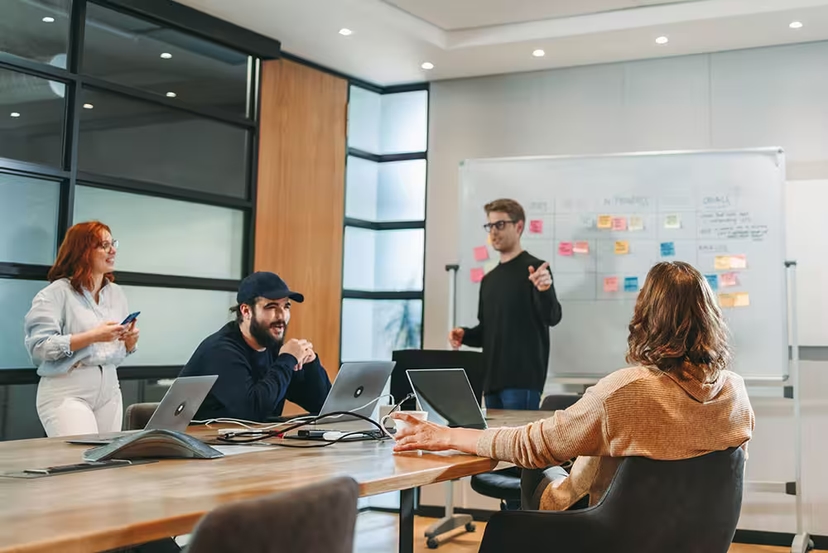 A diverse group of professionals engaged in a team meeting in a modern office, with laptops open and a whiteboard full of notes.