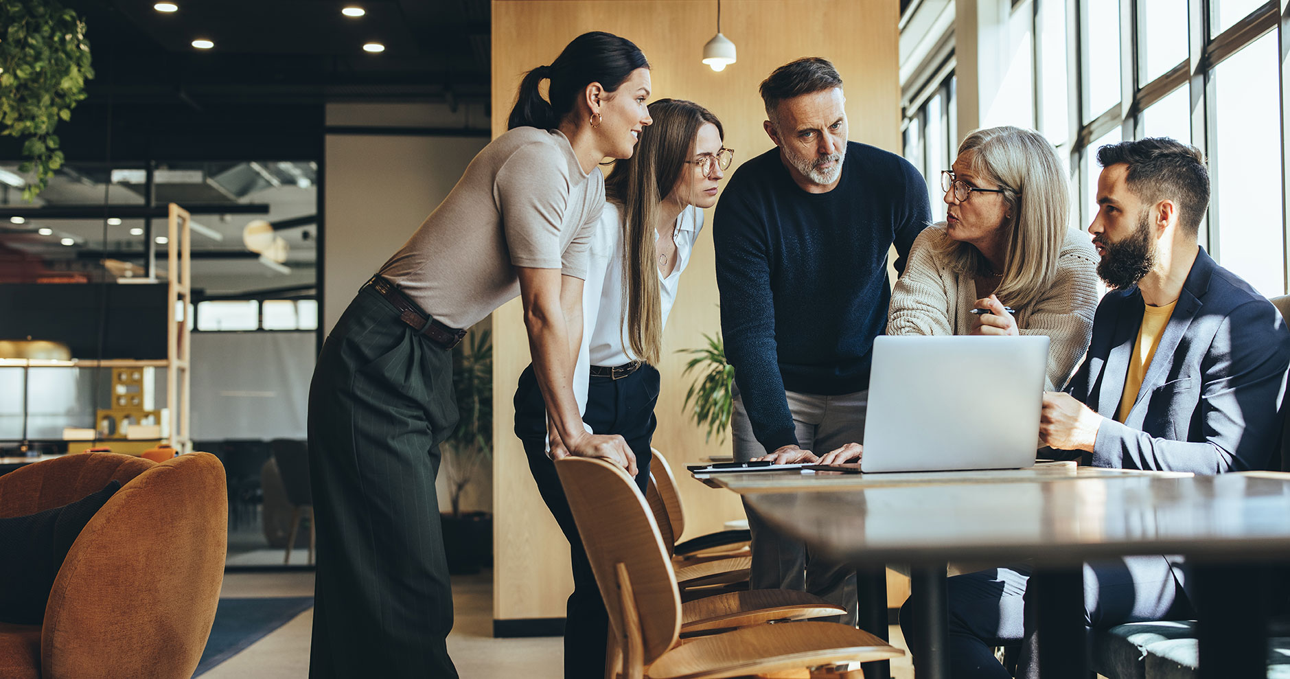 "Group of five people discussing something around a laptop in a modern office setting."